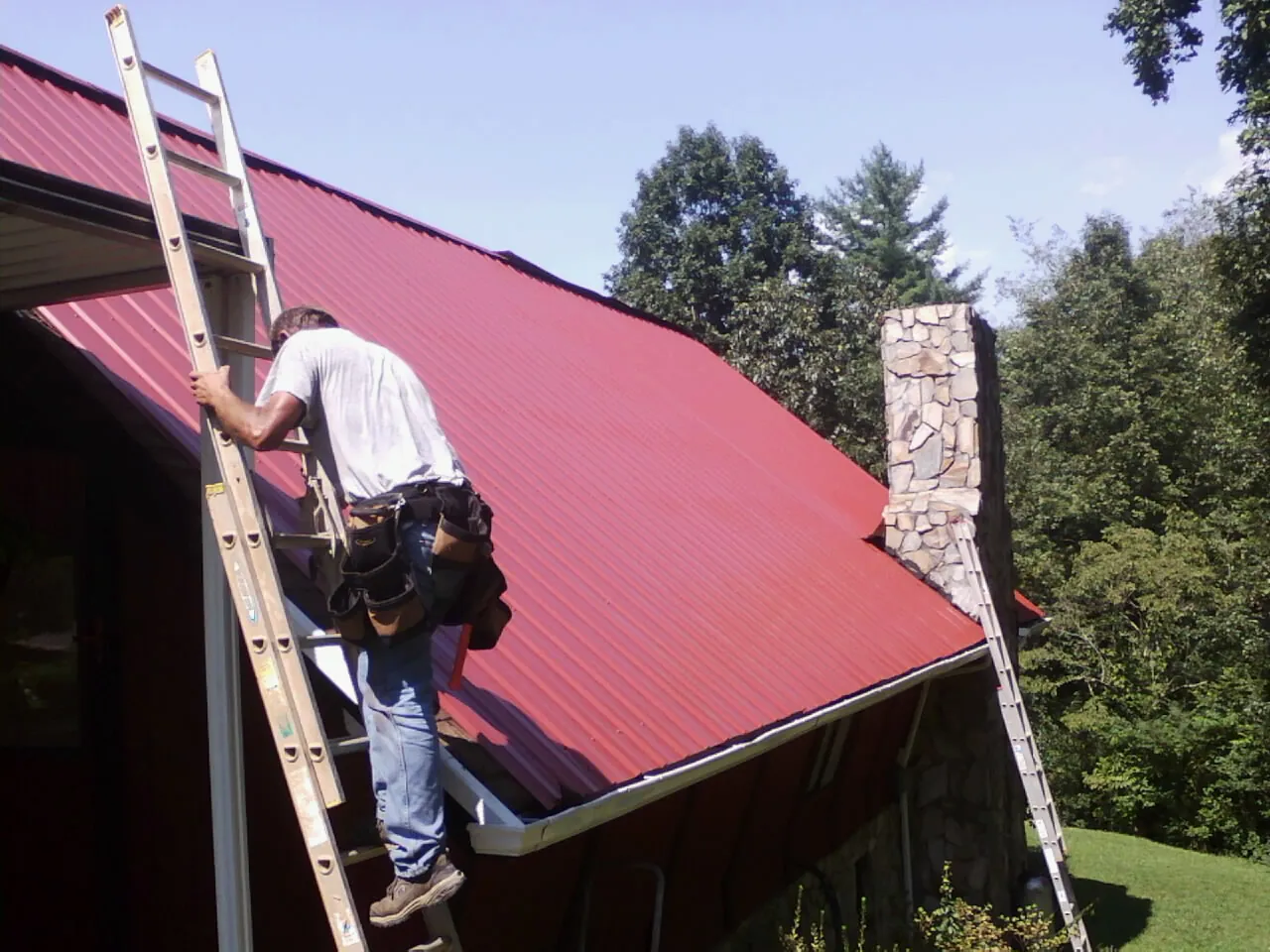 Roofer on a ladder beside a red metal roof and stone chimney — Soffit & Fascia Repair by Roofing Brothers in Lottsburg